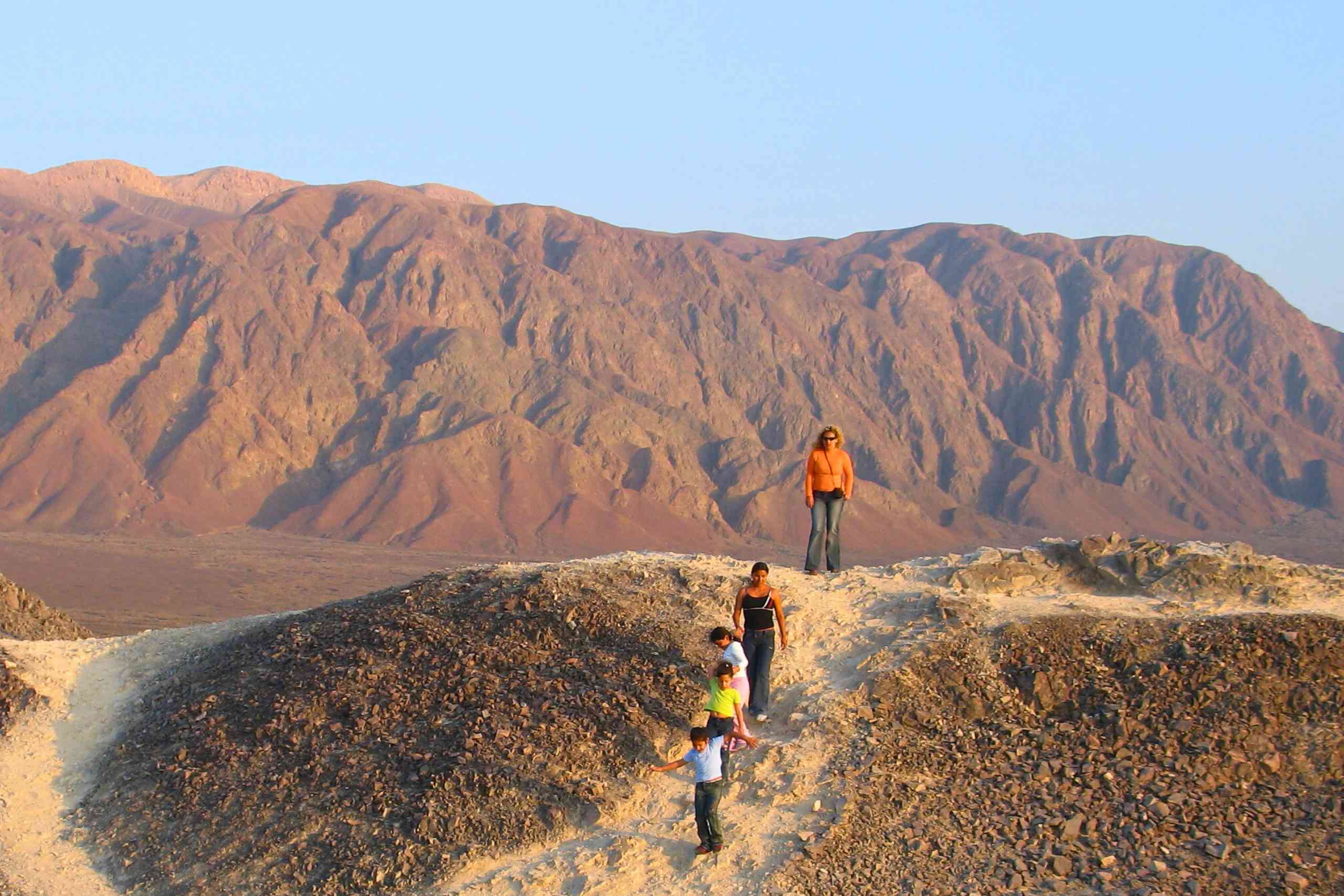 Viajeros caminando por una colina del desierto bajo un cielo azul despejado con montañas rojizas al fondo, cerca de Nazca, en el sur del Perú